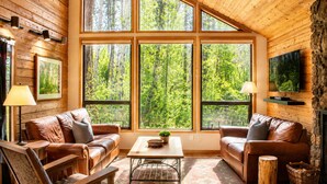 A cozy cabin interior with wooden walls, large windows, leather sofas, a coffee table, and a fireplace.