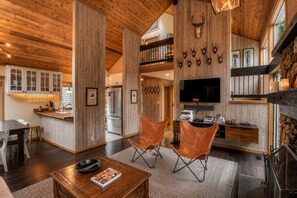 A spacious living area with a wooden ceiling, a fireplace, a television, and a kitchen in the background.