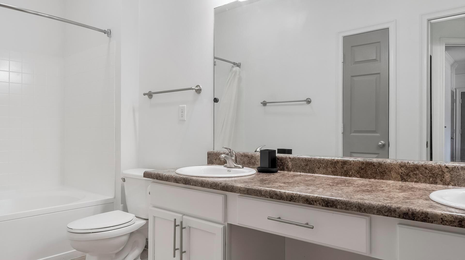 Une salle de bain avec des toilettes blanches, deux lavabos dotés de plans de travail en granit, un grand miroir et un espace douche.