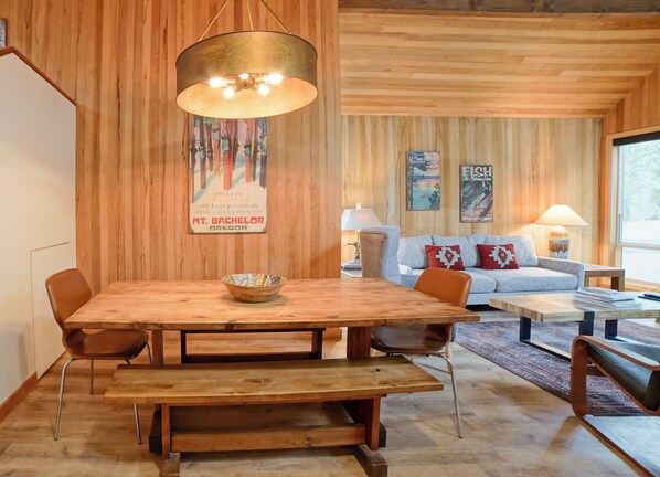 A wooden dining table with a bowl, surrounded by chairs, in a room with wooden walls and a large window.