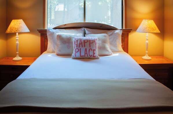 A neatly made bed with a pillow that reads "HAPPY PLACE" and two bedside lamps on wooden nightstands. - Black Butte Ranch House 049 (Black Butte Ranch)