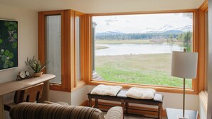 Quarto com uma grande janela que oferece vista para um lago e montanhas. Há um banco de madeira com almofadas, uma mesa com abajur e um relógio na parede.