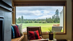 A room with a large window, a wooden bench, a leather chair with a red and black pillow, and a small table with a plant. - Black Butte Ranch Golf Course Condo 081 (Black Butte Ranch)