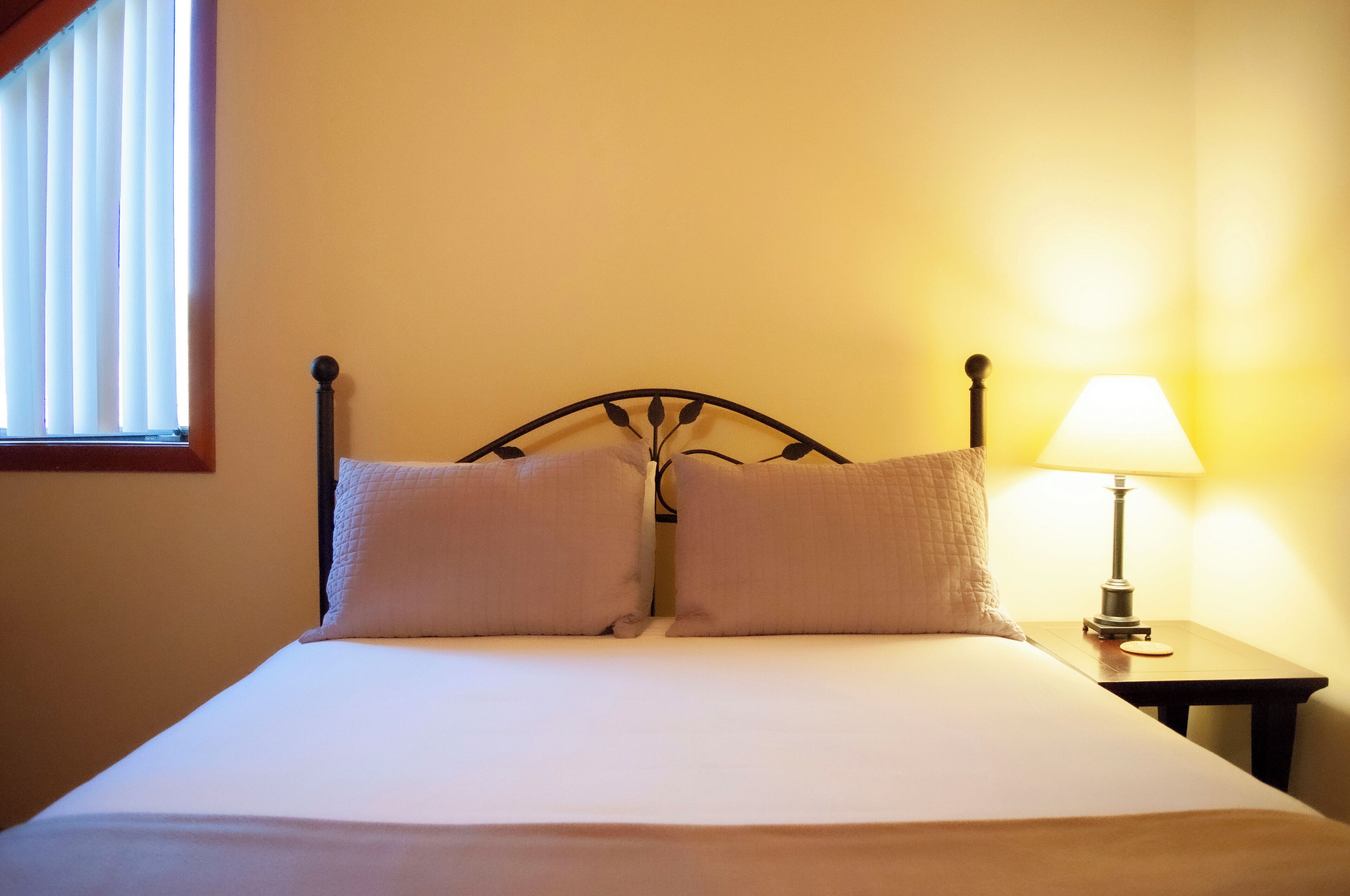 A neatly made bed with two pillows, a bedside lamp, and a window with blinds.
