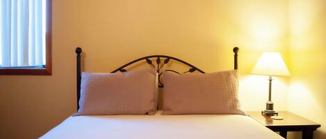 A neatly made bed with two pillows, a bedside lamp, and a window with blinds.