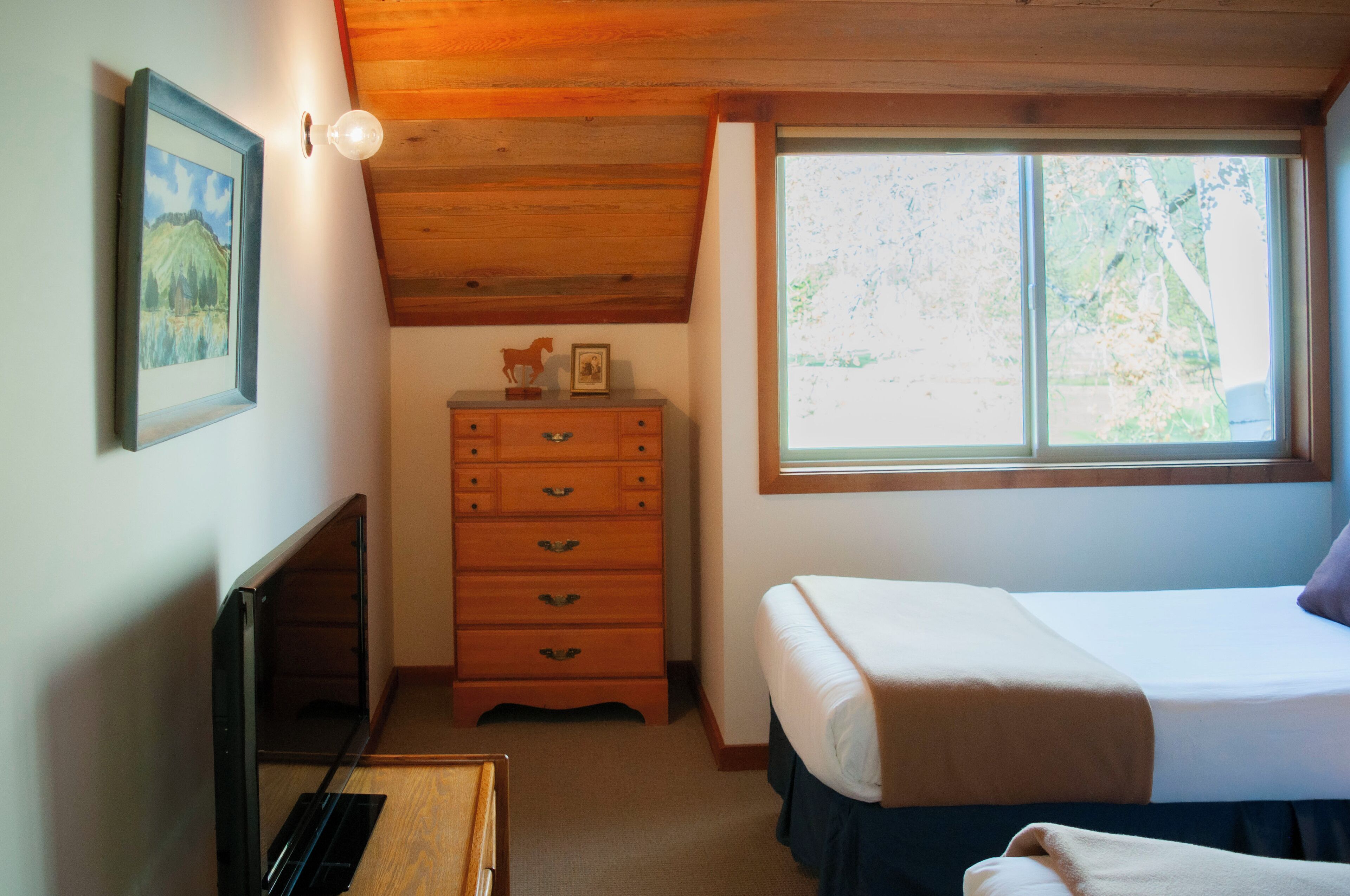 Une chambre à coucher avec un lit, une commode, un tableau encadré au mur et une fenêtre donnant sur des arbres.