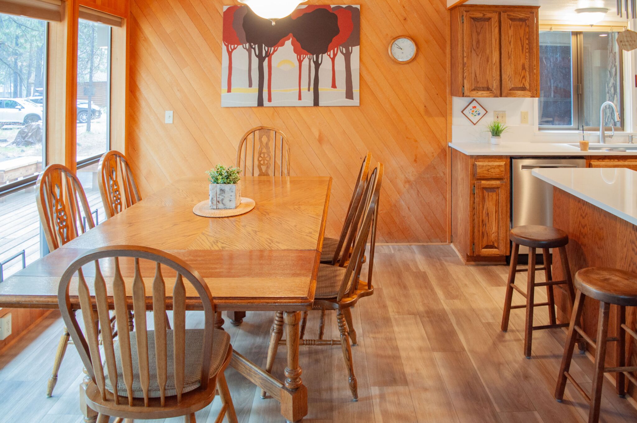 A wooden dining table with chairs, a kitchen area with wooden cabinets, and a large window with a view of the outside.