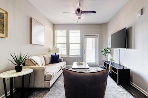 Un salon avec un canapé, une table basse et un téléviseur. On y trouve également un ventilateur de plafond, une plante en pot et une fenêtre avec des stores.