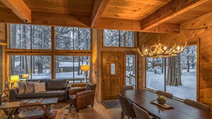 A cozy log cabin interior with a fireplace, large windows, a dining area, and a chandelier. - Black Butte Ranch Golf Home 174 (Black Butte Ranch)