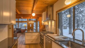 A modern kitchen with wooden beams, a large window with a snowy view, and a central island with a sink and stove. - Black Butte Ranch Golf Home 174 (Black Butte Ranch)