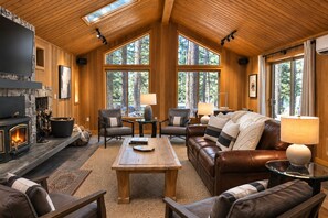 A cozy living room with a fireplace, leather furniture, and large windows offering a view of the forest.