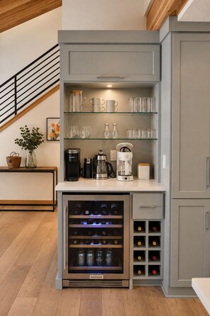 A kitchenette with a built-in wine cooler, a coffee station, and glassware on shelves.