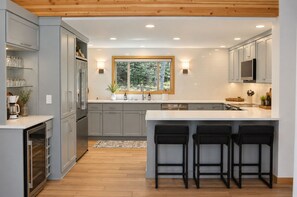 A modern kitchen with grey cabinets, a central island with bar stools, a window with a view of greenery, and recessed ceiling lighting.