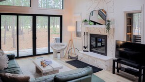 Ein modernes Wohnzimmer mit Kamin aus Stein, einem großen Fernseher und Blick auf den Wald durch raumhohe Fenster.