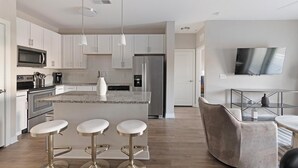Une cuisine moderne avec des meubles blancs, des appareils en acier inoxydable, un îlot central doté de tabourets de bar et un téléviseur à écran plat fixé au mur.