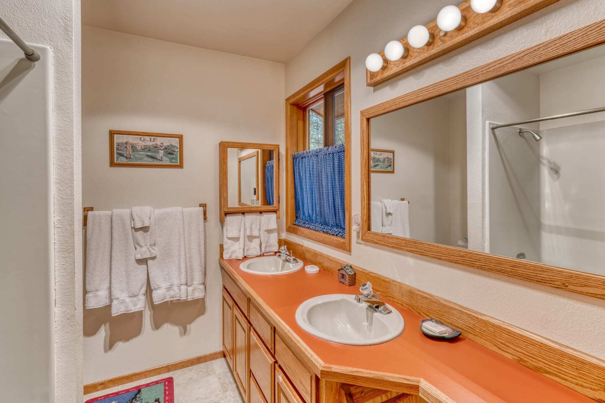 A bathroom with two sinks, wooden cabinets, a large mirror with lights, and towels hanging on a rack.