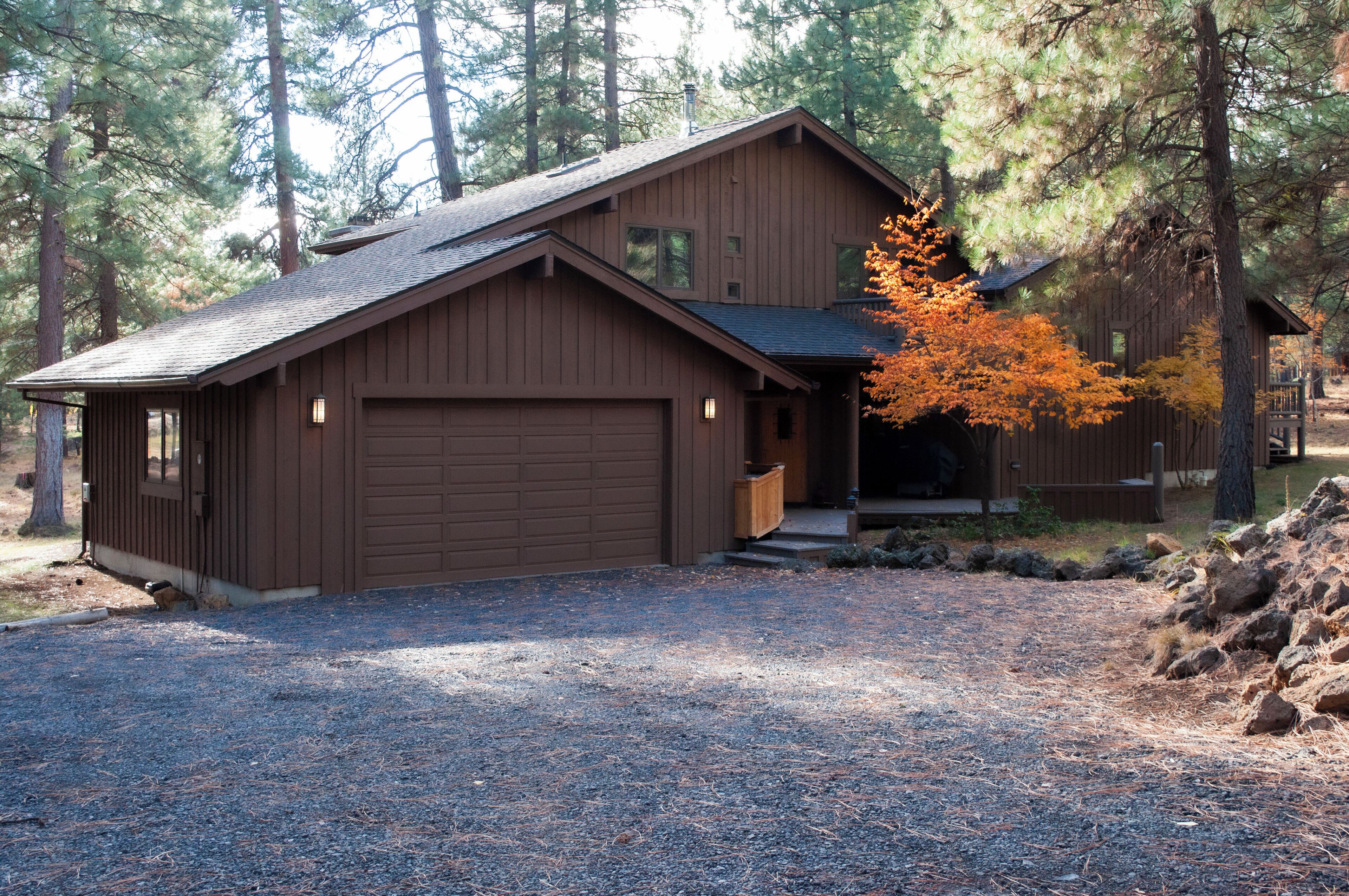 A wooden cabin with a garage, surrounded by trees and a gravel driveway.