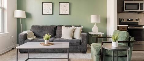 Ein modernes Wohnzimmer mit einem grauen Sofa, zwei gerahmten abstrakten Kunstwerken, einem Couchtisch mit Pflanze und einem grünen Sessel.