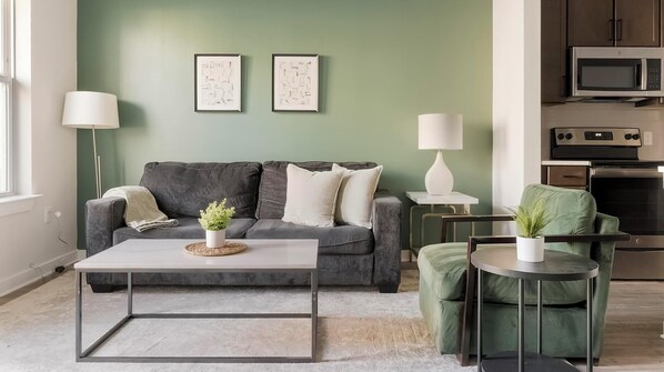 Een moderne woonkamer met een grijze bank, twee ingelijste abstracte kunstwerken, een salontafel met een plant en een groene fauteuil.