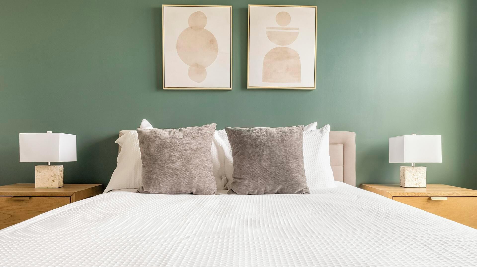 Uma cama bem arrumada com um cobreleito branco texturizado, dois travesseiros cinza e duas mesas de cabeceira com abajures e objetos decorativos.