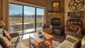 A spacious living room with a stone fireplace, large windows offering a mountain view, and comfortable seating. - Black Butte Ranch Lodge Condo 001 (Black Butte Ranch)