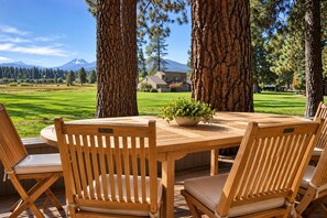 An outdoor wooden table with chairs, surrounded by large trees, with a view of a mountain and a house in the background.