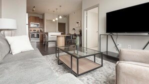 A modern living room with a glass coffee table, a sofa, a TV mounted on a stand, and a kitchen in the background.