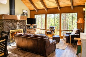 A cozy living room with a stone fireplace, leather sofa, and large windows offering a view of the forest.