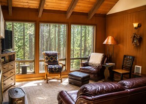 A cozy room with a fireplace, leather furniture, and large windows offering a forest view.