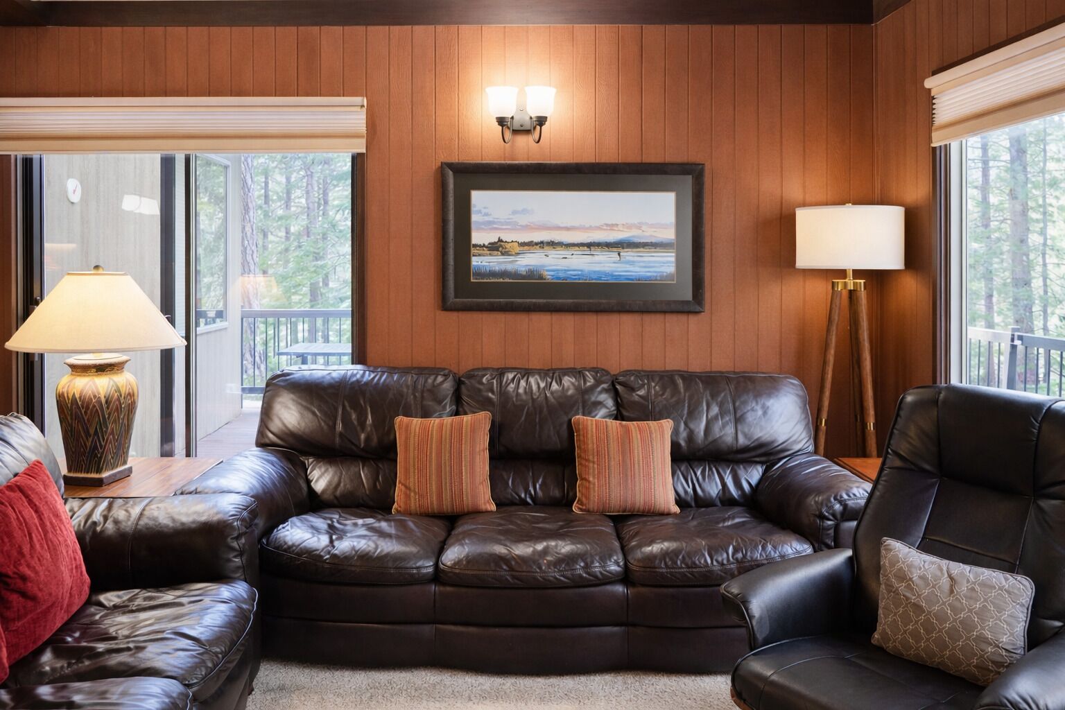 A leather sofa with two cushions, a lamp, a framed picture, and a window with a view of trees.
