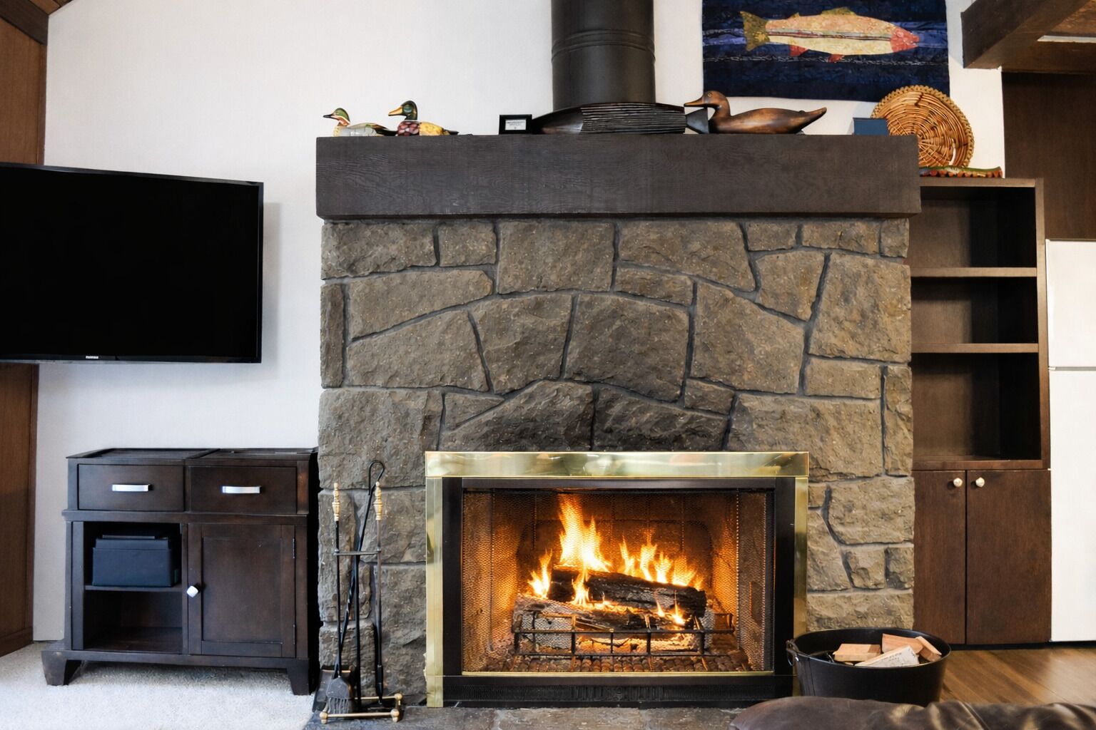 Una chimenea de piedra con fuego, un televisor de pantalla plana, un armario de madera y un estante decorativo con patos.