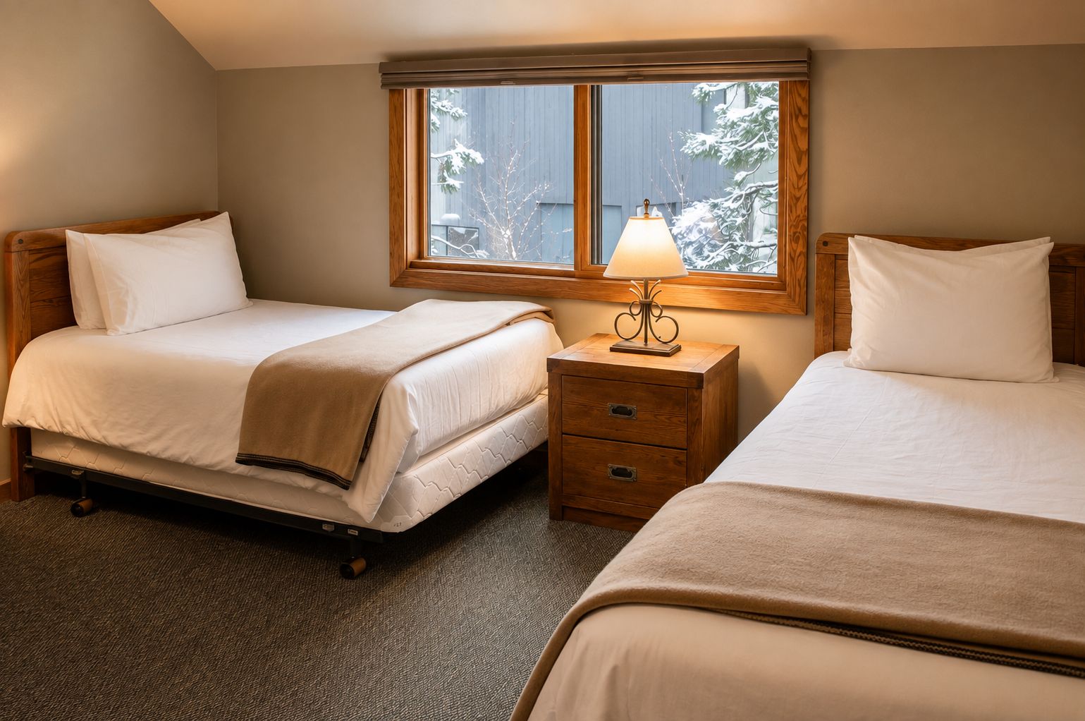 Une chambre d’hôtel avec deux lits, une table de chevet avec une lampe et une fenêtre donnant sur des arbres couverts de neige.