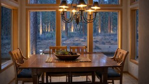 Een eetruimte met een grote houten tafel, zes stoelen, een kroonluchter en uitzicht op een bos door grote ramen.