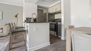 A compact kitchen with a central island, stainless steel appliances, and a dining area with a sofa and chairs. - Landing | Stunning 1BD, Clubhouse, Gym (Winston-Salem)