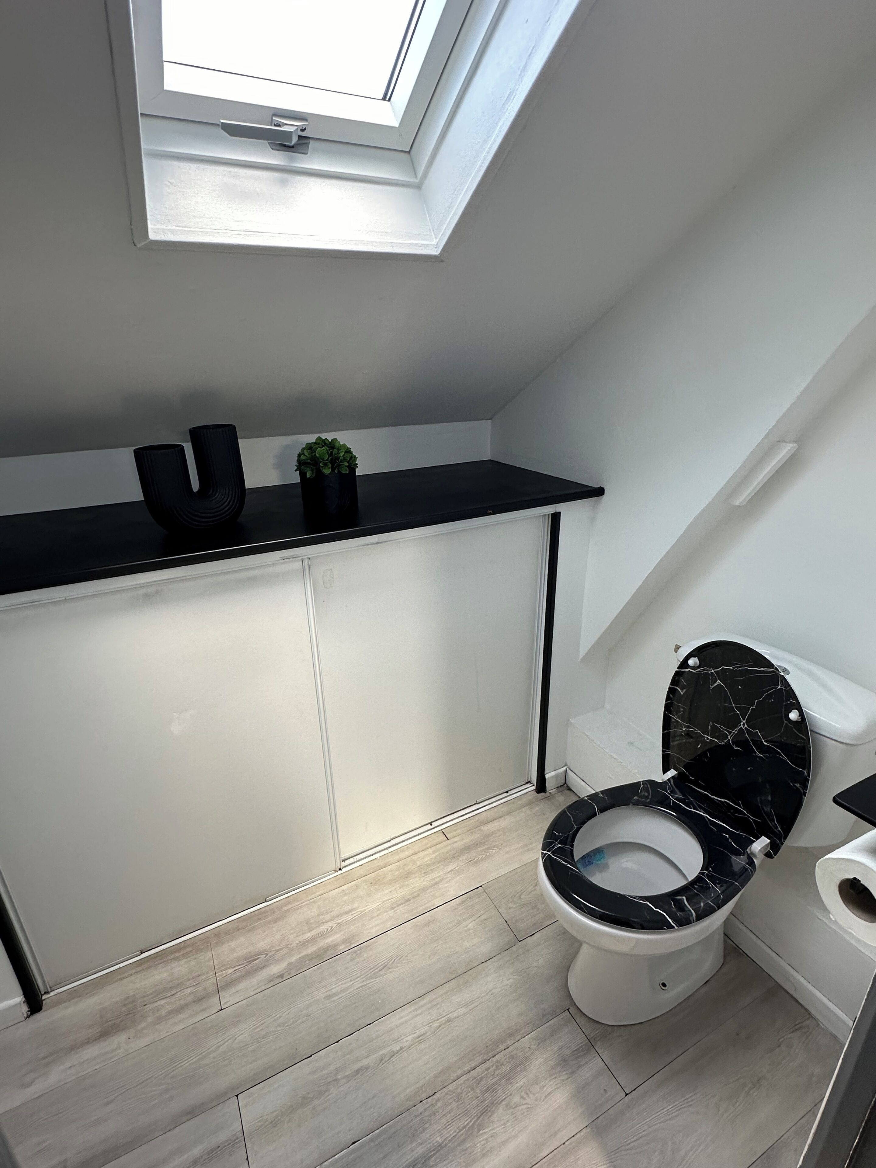 Une salle de bain moderne dotée d’une lucarne, de toilettes noires avec un revêtement en marbre et d’un meuble-lavabo blanc avec un plan de travail noir.