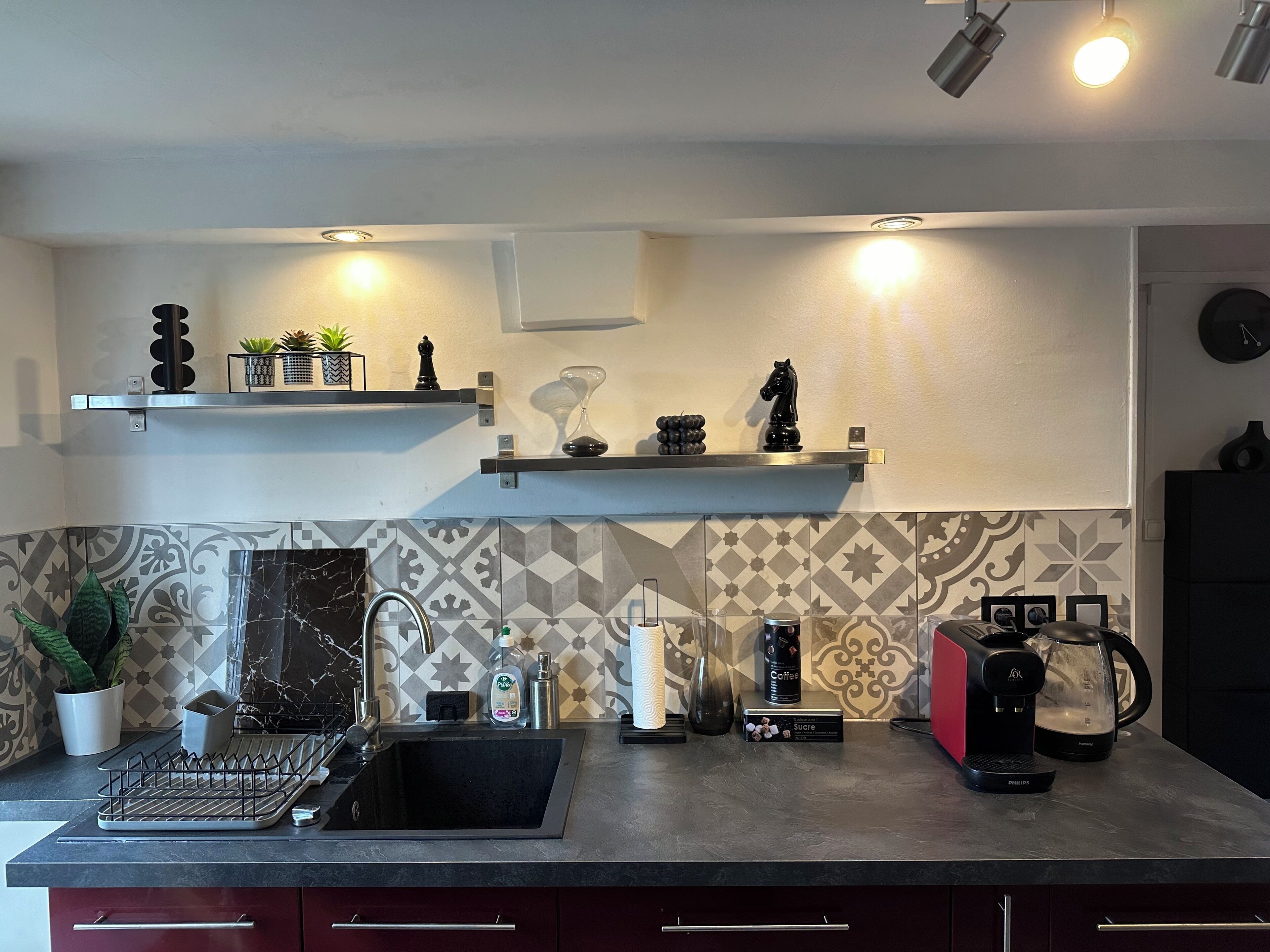 Cozinha moderna com bancada preta, pia de aço inoxidável e diversos eletrodomésticos, incluindo uma cafeteira e uma chaleira. Há objetos decorativos nas prateleiras acima da bancada.