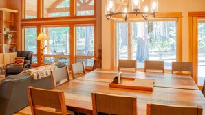A spacious dining area with a large wooden table and chairs, a chandelier, and large windows offering a view of the forest. - Black Butte Ranch South Meadow 119 (Black Butte Ranch)