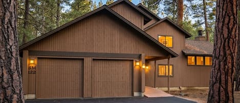 A two-story wooden house with a garage, surrounded by tall trees.