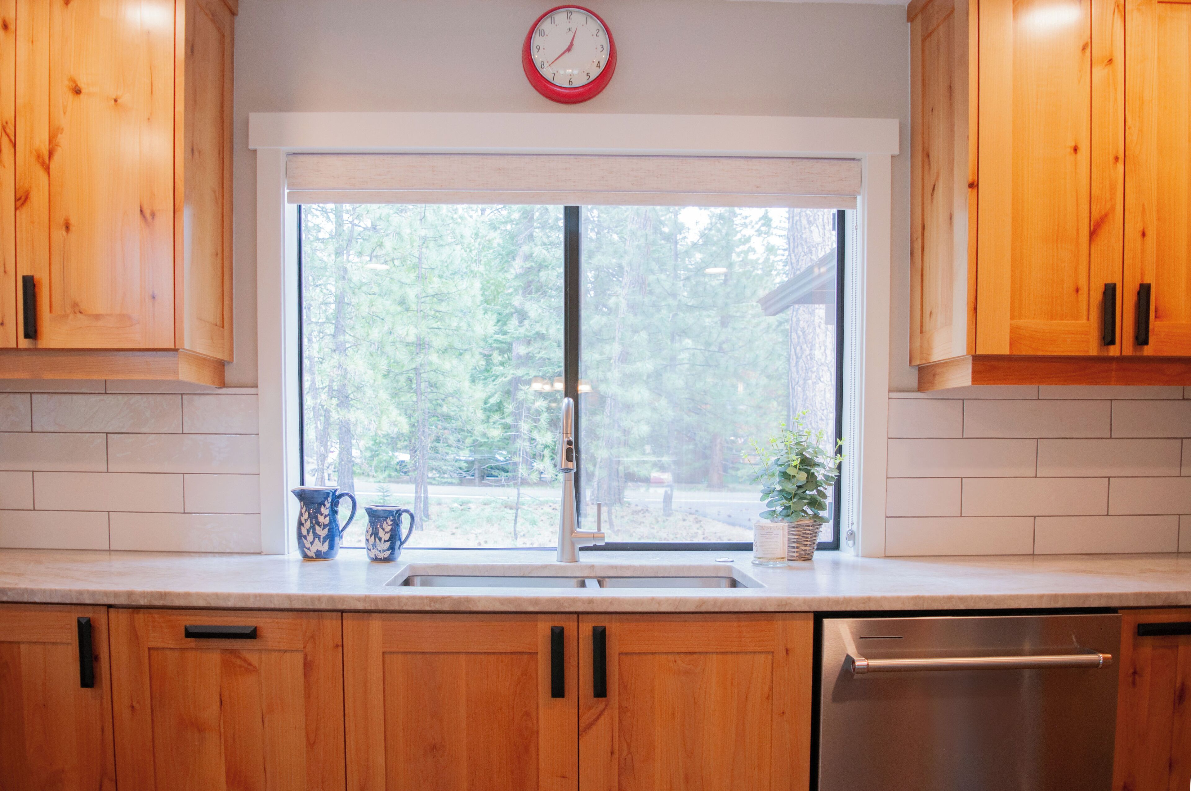 A kitchen with wooden cabinets, a stainless steel dishwasher, and a window with a view of trees.