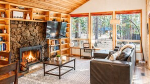 A cozy living room with a stone fireplace, a flat-screen TV, wooden shelves, a sofa, and large windows with a view of trees. - Black Butte Ranch South Meadow 139 (Black Butte Ranch)