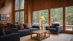 Interior acolhedor de uma cabana com uma grande janela com vista para uma floresta, um sofá, poltronas, uma mesa de centro e um abajur.