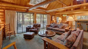 A log cabin interior with a large glass door leading to a deck, furnished with leather chairs and sofas, and a central round table.