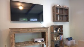 A living room with a wooden TV stand, a shelf with books and decorative items, and a small table with a suitcase. - Renovated 1912 Home 2B/2B w/King&Queen fenced yard (Grants Pass)