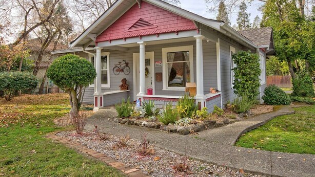 A single-story house with a red roof, a porch with white columns, and a small front yard with a tree and shrubs. - Renovated 1912 Home 2B/2B w/King&Queen fenced yard (Grants Pass)
