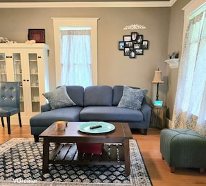 A living room with a blue sofa, a wooden coffee table, a patterned rug, a cabinet, and a wall with framed photos.
