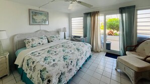 Ein Schlafzimmer mit Bett, Deckenventilator, Nachttisch, Stuhl und Blick ins Freie.