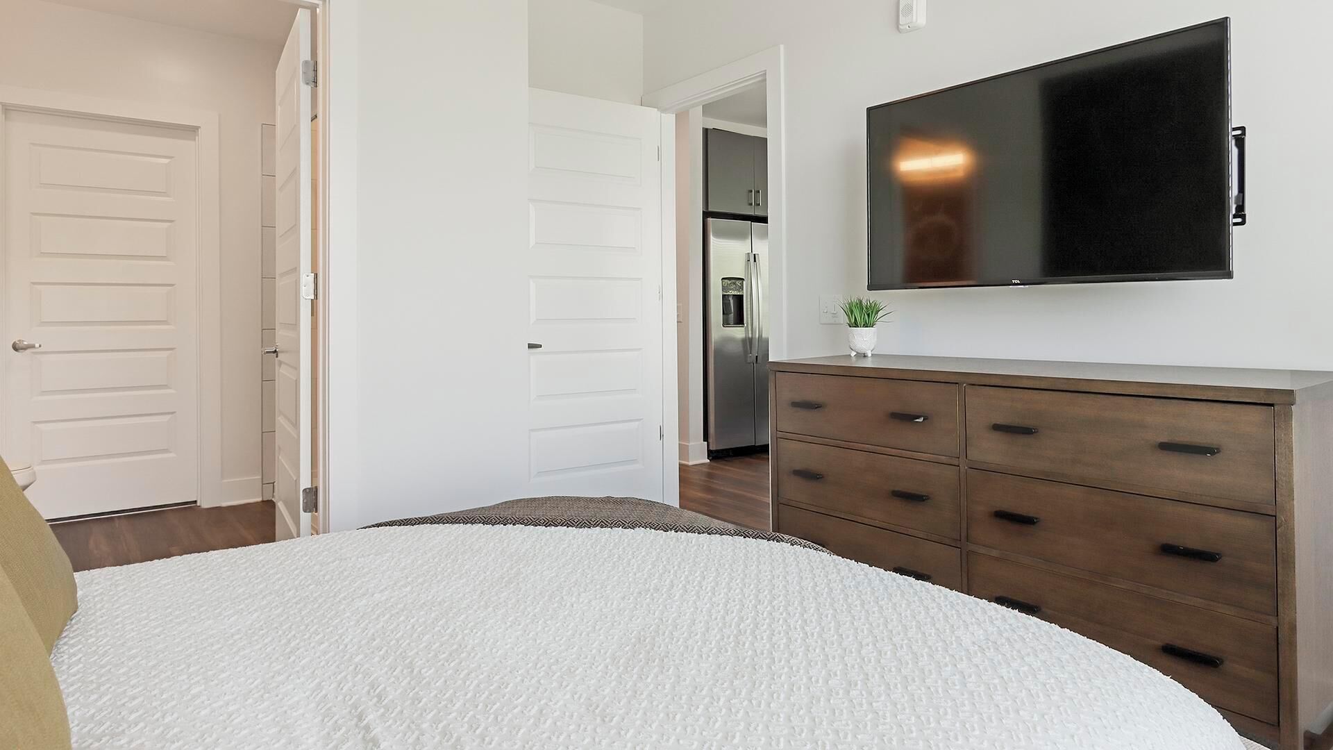A bedroom with a bed, a dresser, a TV, and a door leading to a kitchen.