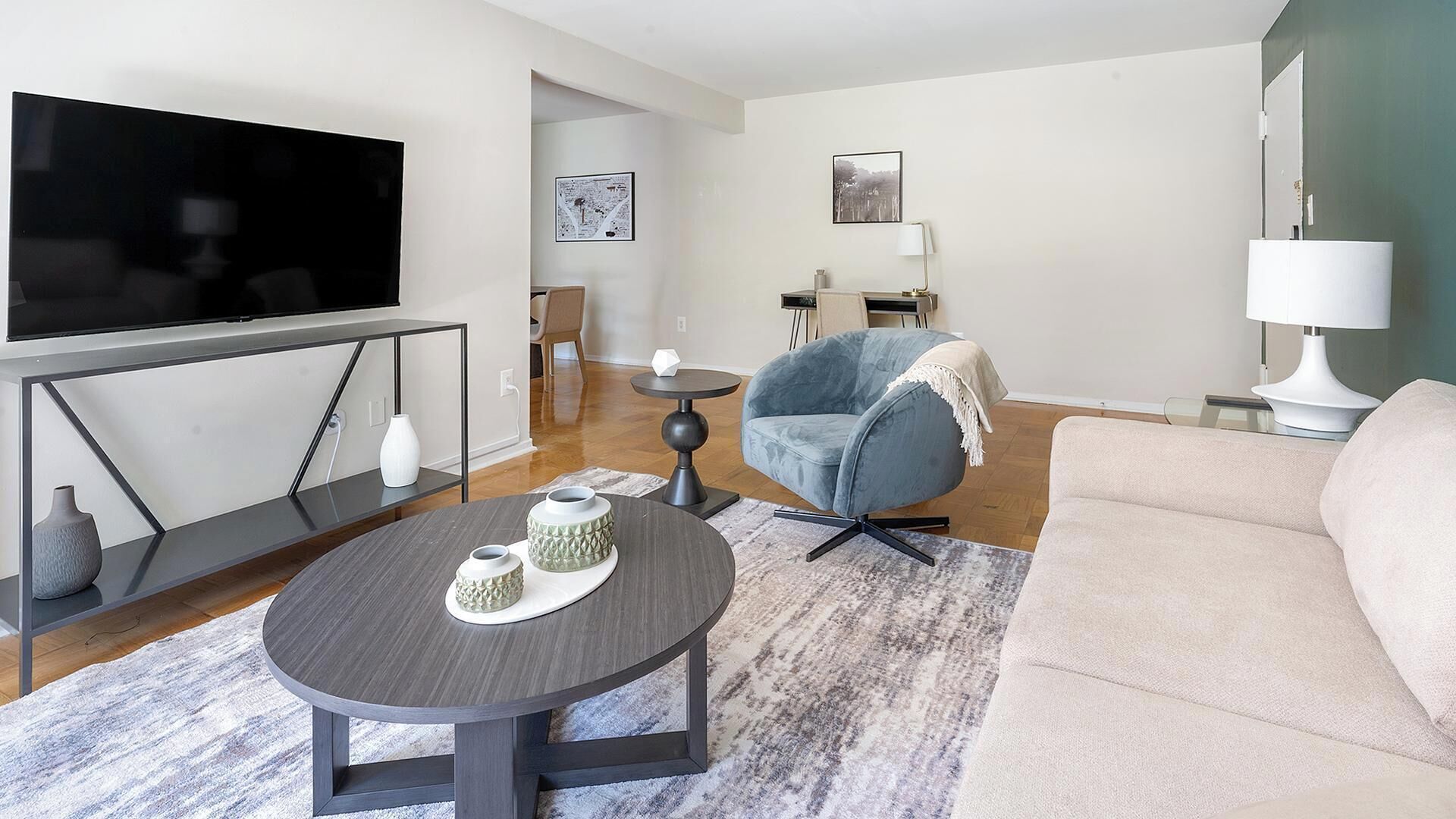 A modern living room with a flat-screen TV, a round coffee table with decorative items, a blue armchair, a beige sofa, and a small side table with a lamp.