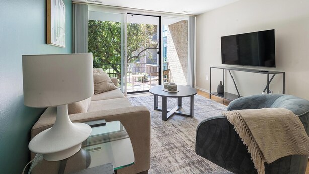 Ein Wohnzimmer mit einer Couch, einem Tisch, einer Lampe, einem Fernseher und Blick ins Freie durch eine Glastür.