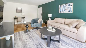 Ein modernes Wohnzimmer mit einem beigefarbenen Sofa, einem blauen Sessel und einem runden Couchtisch. An der Wand hängt ein Gemälde und auf einem Beistelltisch steht eine Lampe.
